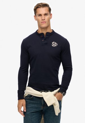 Navy cotton long-sleeve henley shirt with a three-button placket, embroidered logo on the chest, worn with light-colored sweater tied around the waist.
