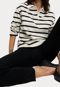 Personne assise avec une jambe pliée, portant une chemise à manches longues rayée noire et blanche, un pantalon noir et des chaussures noires à enfiler.