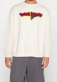 Una persona indossa un felpa off-white con un design colorato "Wow Lights" sul petto e pantaloni grigi, in piedi contro uno sfondo semplice.