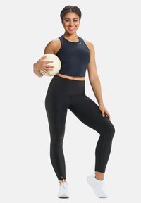 Musta crop top ja korkeavyötäröiset mustat legginssit, joiden pinta on sileä. Pidessä vaaleaa lentopalloa. Valkoiset urheilu kengät.