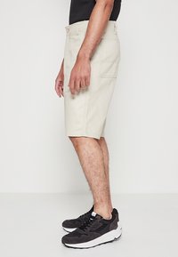 Bermudas beige hasta la rodilla, hechas de una tela texturizada, cuentan con bolsillos laterales y un cierre de botón. Combinadas con zapatillas deportivas negras.