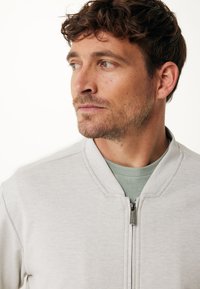 Světlé šedé bomber bundy s texturovaným pletením, zapnutým zipem a kulatým výstřihem. Nosené přes světle zelené tričko.