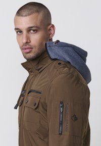 Braune Jacke mit einer grauen Fleecekapuze, mehreren Reißverschluss- und Druckknopftaschen sowie einem Logodetail am Ärmel. Das Material hat eine glatte, strukturierte Oberfläche.