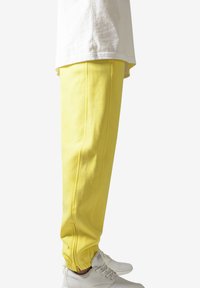 Pantaloni de trening galbeni cu o textură netedă, având buzunare laterale, un design cu piciorusuri conice și manșete elastice. Asortați cu adidași albi.