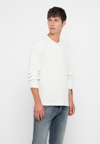 Joven vestido con una camiseta de punto waffle blanca de manga larga con botones y vaqueros azules desgastados, de pie contra un fondo blanco liso.