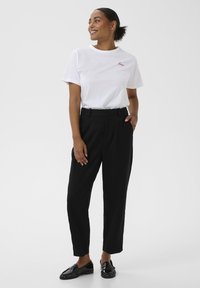 Femme debout, souriante, portant un T-shirt blanc avec le texte "Love", un pantalon noir taille haute et des mocassins noirs, sur fond clair uni.
