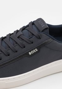 Marinblå sneaker med slät syntetisk ovandel, texturerad tygtunga, vit gummisula och framträdande "BOSS"-logotyp på sidan.