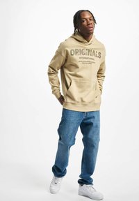 Beige hoodie with "ORIGINALS" text, front pocket, paired with blue jeans and white sneakers. Casual fit, soft fabric, minimal design.