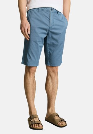 Homme portant un short bleu au genou, un T-shirt blanc et des sandales marron à double boucle, debout les mains dans les poches devant un fond blanc.