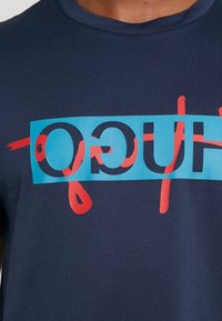 Maglietta in cotone blu navy con un audace design grafico. Rettangolo centrale in teal con testo rosso e nero, accentuato da gocce rosse. Maniche corte.