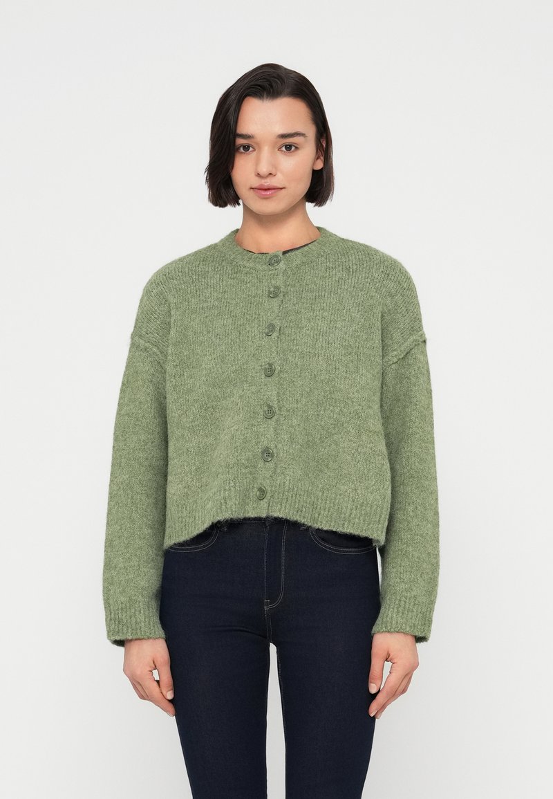 Πράσινη πλεκτή ζακέτα με στρογγυλή λαιμόκοψη, κούμπωμα με κουμπιά και μακριά μανίκια. Διαθέτει κοντό σχέδιο και ριμπ γύρω από το τελείωμα.