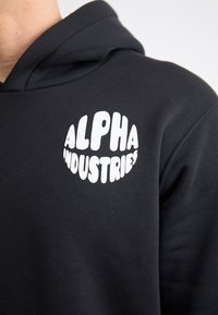 Moletom preto com o logo branco "ALPHA INDUSTRIES", apresentando uma textura de tecido macio e um corte descontraído. Sem padrões ou detalhes adicionais.