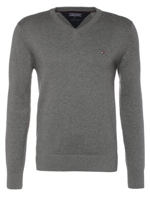 Uomo che indossa un maglione grigio a scollo a V, jeans scuri, sneakers nere, che tiene una tracolla di uno zaino blu navy sulla spalla, camminando in avanti.