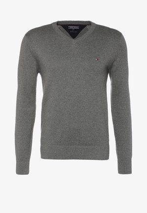 Mann trägt grauen V-Neck-Pullover, dunkle Jeans, schwarze Sneakers, hält einen marineblauen Rucksackriemen über der Schulter und geht vorwärts.