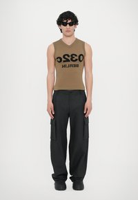 Ärmelloses braunes Tanktop mit der Aufschrift "032 BERLIN" in schwarzer Schrift, kombiniert mit locker sitzenden schwarzen Cargo-Hosen mit Seitentaschen.