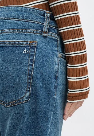 Sinised teksapüksid tuhmunud viimistlusega, millel on oranžid õmblused ja väike tikitud logo tagataskul. Triibuline pruun pika varrukaga topp.