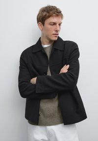 Sorte uldjakke med krave, frontknaplukning og tofarvet overdesign. Båret over en beige striktrøje mod en hvid baggrund.