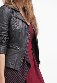 Veste de moto en cuir noir avec fermeture éclair asymétrique, éléments en argent et manches texturées, superposée à une robe bordeaux à rayures.