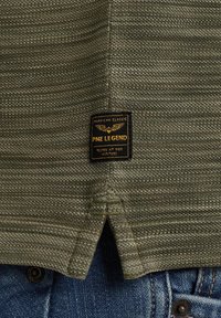 Chemise tricotée vert olive avec une étiquette indiquant "PME Legend" en noir et or. Présente un ourlet fendu en bas et un tissu texturé.