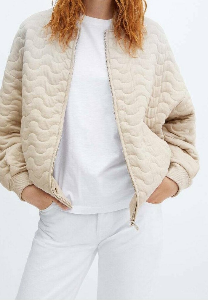 Beige gesteppte Bomberjacke mit welliger Textur, gerippten Bündchen und Saum sowie einem Reißverschluss vorne, getragen über einem weißen T-Shirt und weißen Hosen.
