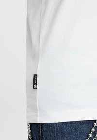 T-shirt bianco in cotone con taglio dritto, dotato di un'etichetta laterale nera con testo bianco. Jeans in denim texturizzato con accentuazione a catena.