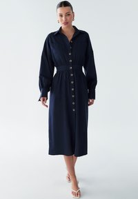 Vestito camicia blu navy realizzato in tessuto leggero, caratterizzato da maniche lunghe, cintura in vita e dettagli dorati sui bottoni.