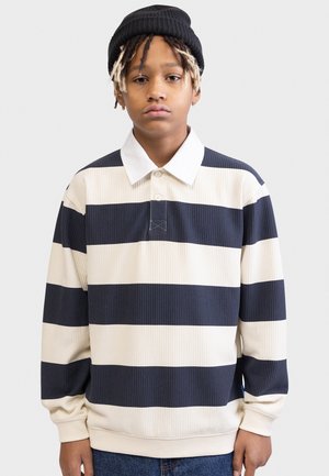 Młoda osoba z krótkimi dredami, nosząca czarną czapkę typu beanie oraz granatowo-białą koszulę rugby w szerokie paski, stoi na jednolitym tle.