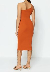Robe orange à une épaule, côtelée, ajustée et arrivant au genou. Associée à des sandales dorées à lanières avec des accents floraux près des orteils.
