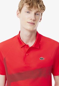 Röd polo-tröja tillverkad av mjuk tyg, med krage, tvåknaps placket och Lacoste-logotyp i grönt på vänstra bröstet.