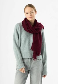 Sciarpa bordeaux con bordi sfrangiati appoggiata su un maglione grigio chiaro. Il maglione ha un orlo a coste e una vestibilità leggermente ampia. Jeans grigio chiaro.