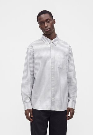 Homme portant une chemise à rayures verticales grises et blanches à manches longues et un pantalon noir, debout devant un fond blanc uni.