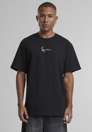 Férfi fekete, túlméretezett pólóban, amelyen fehér "Kani" feliratú logó található az elején, egy egyszerű szürke háttér előtt áll.