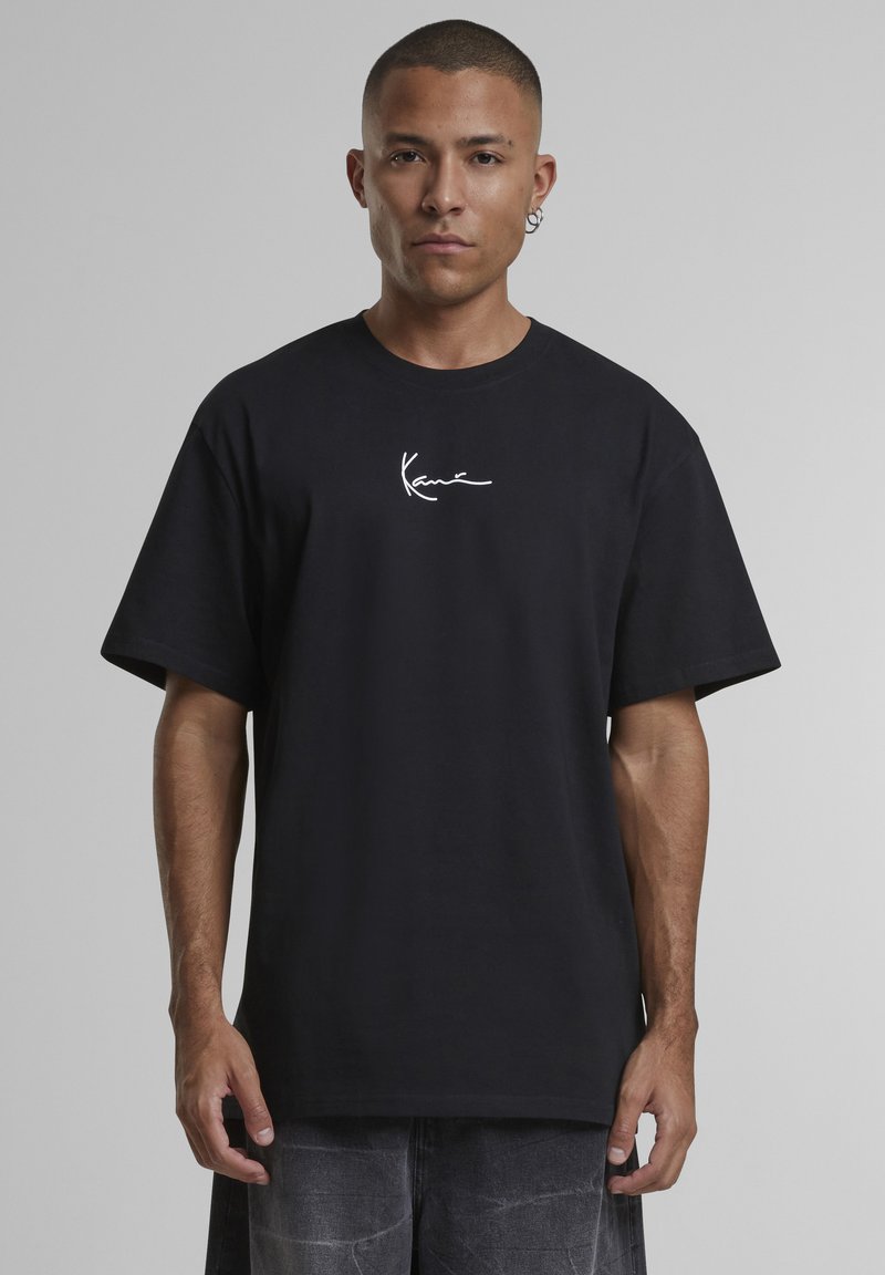 Άντρας που φοράει μαύρο φαρδύ T-shirt με λευκό λογότυπο "Kani" μπροστά, στέκεται μπροστά σε απλό γκρι φόντο.