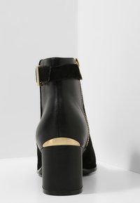 Bota de cuero negro hasta el tobillo con una correa de ante en el tobillo, detalles en oro y un tacón bloque moderado. Cuenta con una cremallera lateral para facilitar su uso.