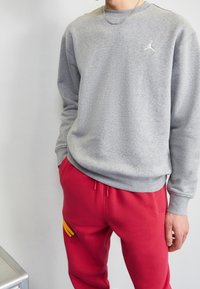 Persona vestida con un sudadera gris con un logo blanco de Jordan y pantalones deportivos rojos con un gráfico naranja en el muslo izquierdo, con las manos en los bolsillos.
