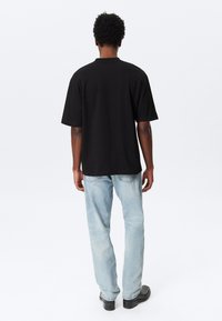 Person som står med ryggen til iført en svart kortermet t-skjorte, lyseblå jeans og svarte sko mot en ensfarget hvit bakgrunn.