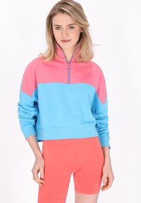 Sudadera con cremallera de bloques de color en rosa y azul, con mangas largas y puños de canalé, combinada con pantalones cortos ajustados en coral. Textura de tela suave.