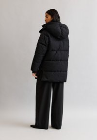Schwarze Pufferjacke mit Kapuze, ausgestattet mit genähten Paneelen und einem etwas längeren Rückenteil. Modell trägt weite schwarze Hosen und steht vor einem hellen Hintergrund.