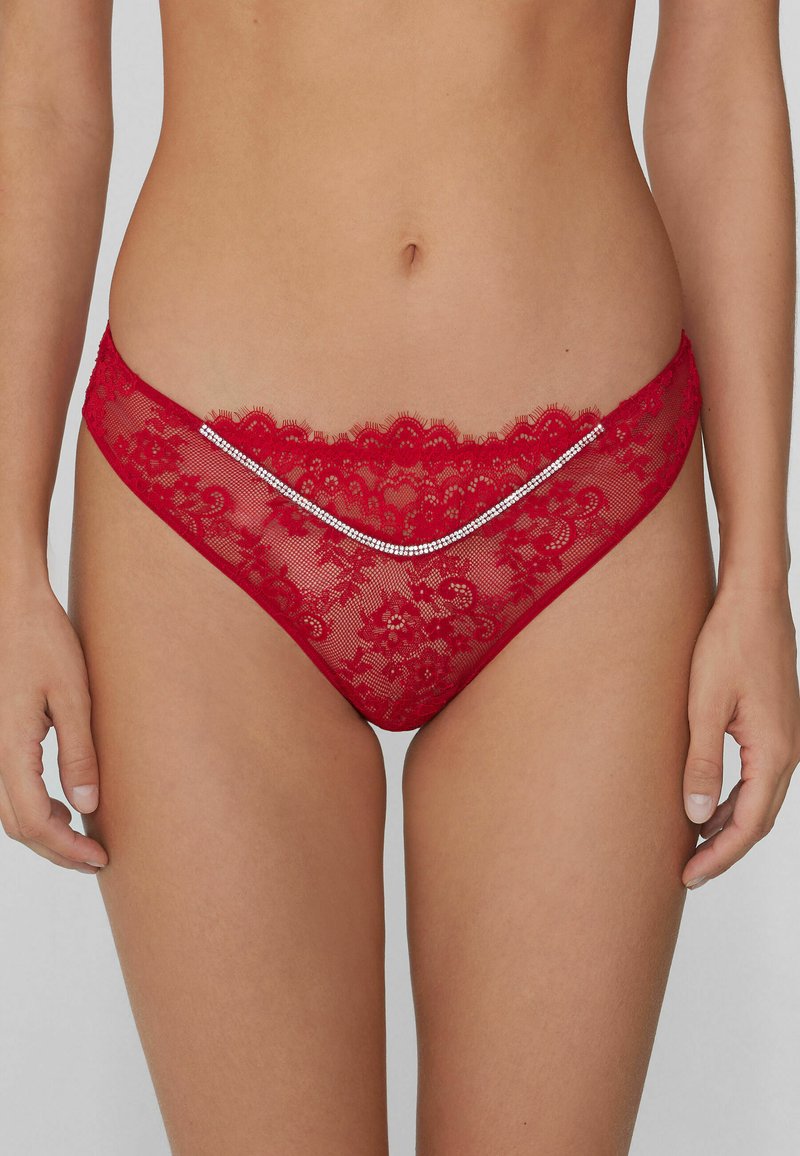 Red lace thong featuring floral patterns, scalloped edges, and a silver rhinestone accent across the front. Sheer texture and low-rise design.