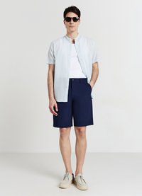 Hellblaues, kurzärmeliges Hemd mit Knöpfen, weißes Tanktop, dunkelblaue Shorts und hellgraue Schuhe. Sonnenbrille vervollständigt das Outfit.