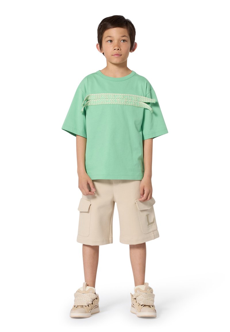 T-shirt surdimensionné vert menthe avec un accent tressé, associé à un short cargo beige avec poches, et des baskets de couleur crème avec des détails texturés.