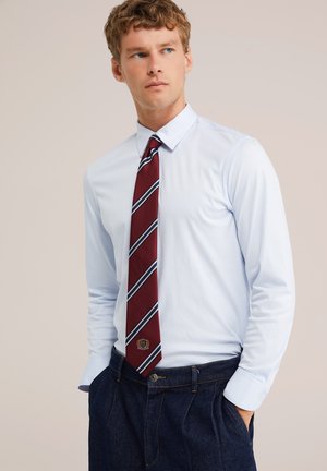 Jeune homme portant une chemise bleu clair, une cravate bordeaux rayée et un jean bleu foncé, regardant légèrement sur le côté, les mains dans les poches.