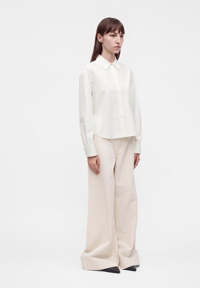 Femme debout portant une chemise blanche boutonnée à manches longues et un pantalon large beige clair couvrant des chaussures noires pointues.