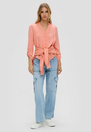 Pfirsichfarbene Bluse mit Knopfleiste und dreiviertel Ärmeln, tailliert mit einem Knoten an der Taille, kombiniert mit hellblauen Cargo-Jeans und weißen Sneakern.