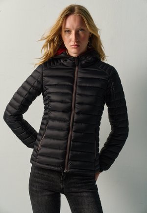 Mujer rubia que lleva una chaqueta negra acolchada con cremallera y vaqueros oscuros, de pie con las manos en las caderas contra un fondo claro y liso.