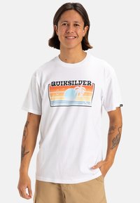 T-shirt bianco in cotone con una grafica colorata che presenta un tramonto, una palma e onde. Maniche corte e scollatura rotonda.