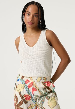 Haut blanc sans manches en maille avec un col en V. Présente un tissu texturé et une silhouette ajustée. Associé à un pantalon coloré à motifs.