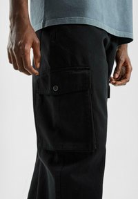 Man in zwarte cargobroek met grote zijzak en drukknoop, zichtbaar van de taille tot halverwege de dijen met handen ontspannen aan de zijkanten.