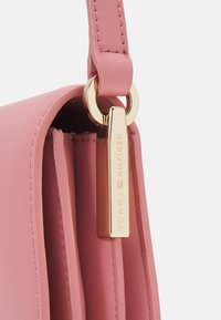 Rosa Lederhandtasche mit glatter Textur, versehen mit einem vergoldeten Hardware-Tag, das mit "TOMMY HILFIGER" geprägt ist, und einem passenden Trageriemen.