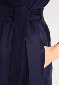 Námořnické zavinovací šaty s látkovým páskem, obsahující boční kapsu. Hladká textura a čisté linie zvýrazňují minimalistický design.